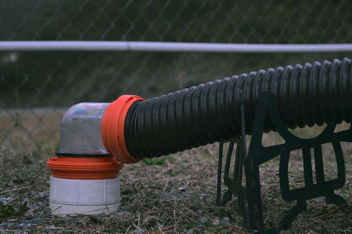 A close-up of an RV sewer hose connected to a dump station using a transparent elbow fitting and an orange sewer hose adapter. The hose is supported by a black plastic sewer hose support, ensuring a proper slope for efficient drainage. The setup prevents tangling and keeps the system clean and organized.