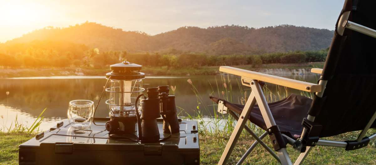 A black folding camping chair positioned by a peaceful lakeside at sunset, with a lantern, binoculars, and a glass on a storage box nearby. The scene highlights the comfort and relaxation that zero gravity chairs bring to an outdoor RV experience.