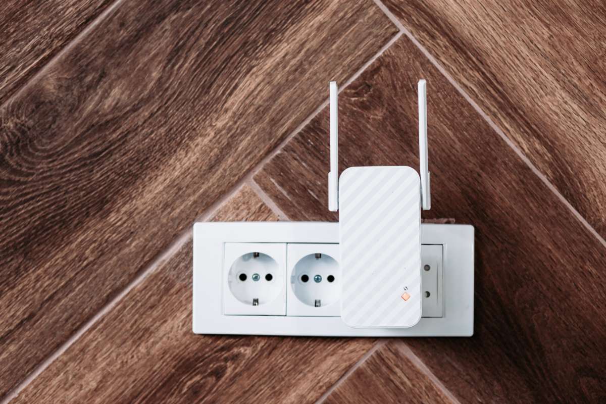 A white Wi-Fi range extender with two antennas plugged into a dual European power socket on a wooden wall. The device enhances wireless connectivity, similar to how a cell phone signal booster ensures reliable communication in remote areas.