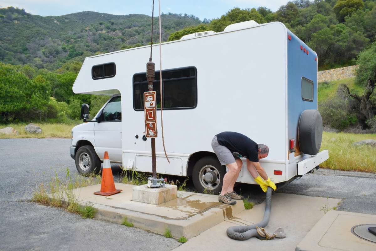 A person emptying the wastewater tank of an RV at a dump station, wearing yellow gloves and using a hose connected to the black water outlet. The image highlights the importance of using a transparent sewer tank rinser to ensure an efficient and thorough cleaning of the RV's sewer system.