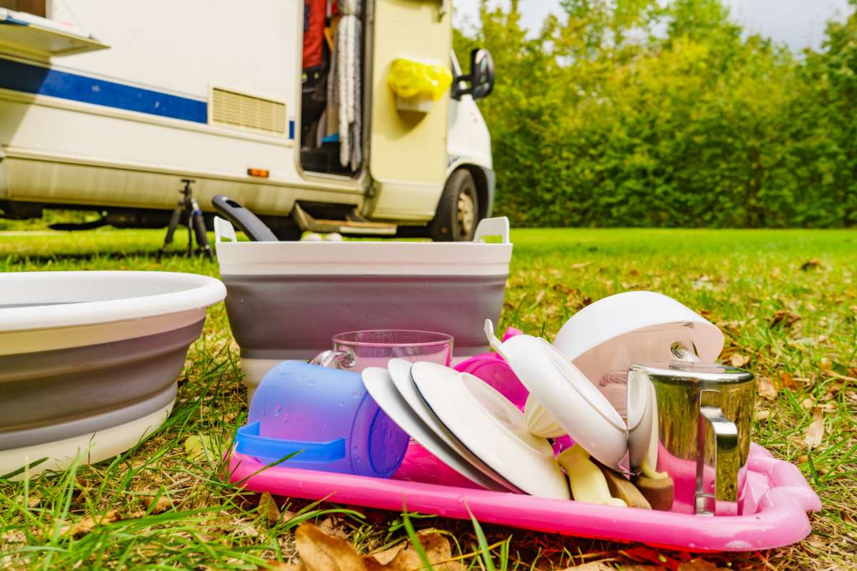 A colorful assortment of washed dishes, cups, and utensils placed on a pink drying tray on the grass next to a camper van. Two collapsible dishwashing tubs are also visible, emphasizing space-saving and practical solutions for RV kitchens.