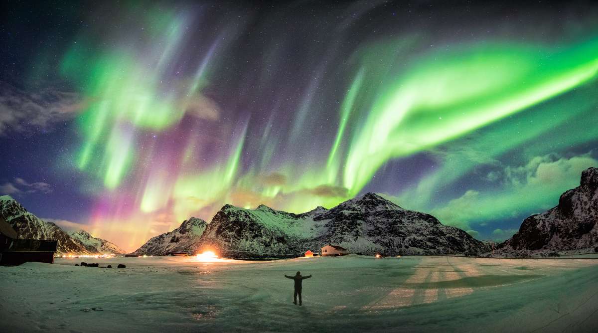 A person standing in a snowy landscape under the vibrant Northern Lights, with mountains and small houses in the background. The image captures the magical winter experience in Iceland, highlighting the seasonal differences that affect RV travel, such as daylight hours and road accessibility.
