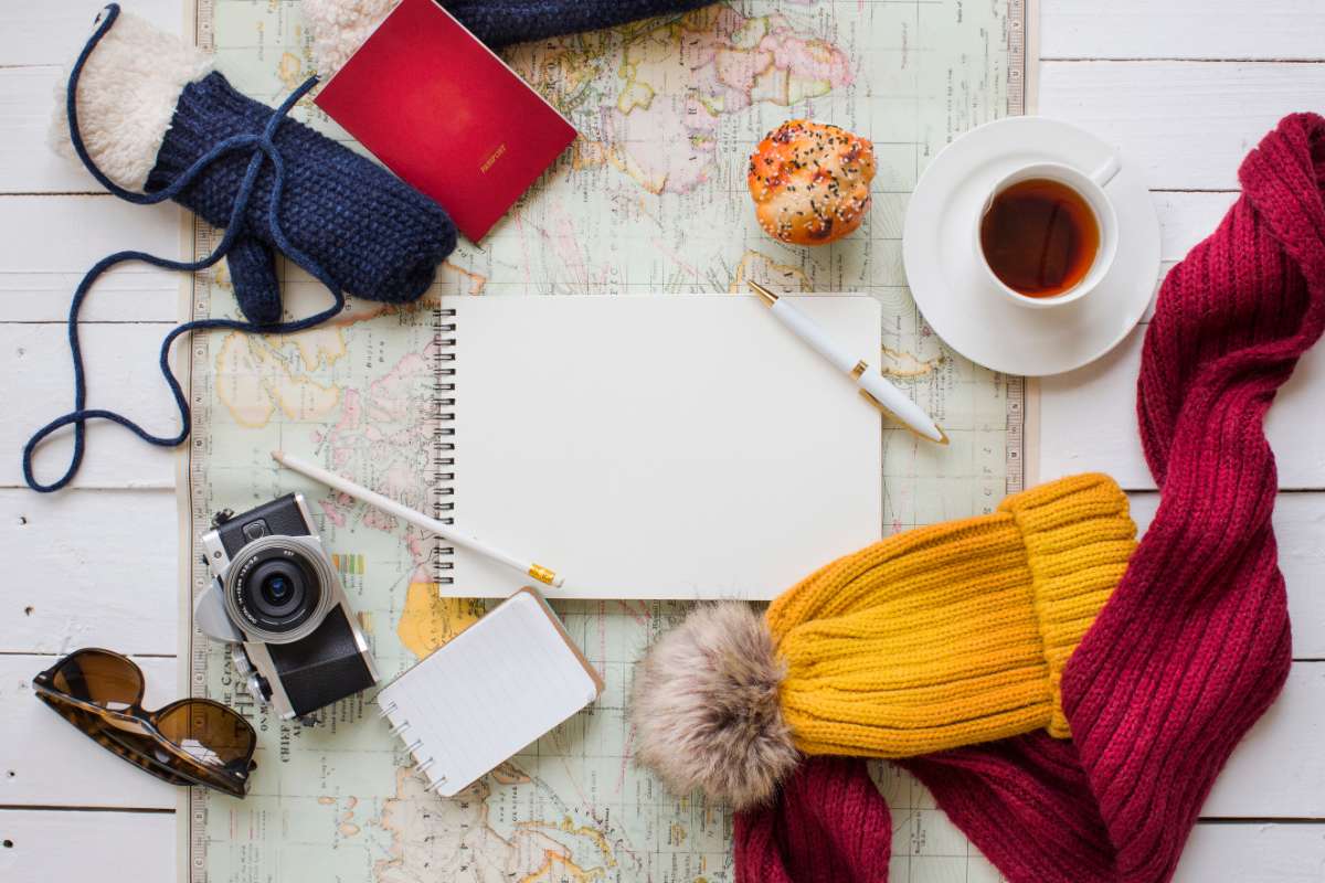 A flat lay of travel essentials on a world map, including a notebook, warm gloves, a yellow beanie, a red scarf, a passport, a vintage camera, sunglasses, a cup of tea, and a muffin. The image represents planning and packing for an RV trip in Iceland, emphasizing the importance of warm clothing and essential travel gear.