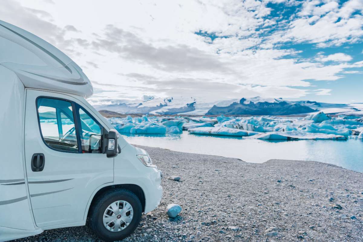 A white RV parked on a rocky shoreline in Iceland, overlooking a glacial lagoon with floating icebergs and snow-capped mountains in the background. The image highlights the freedom and adventure of exploring Iceland by campervan, offering flexibility and cost savings on accommodation.