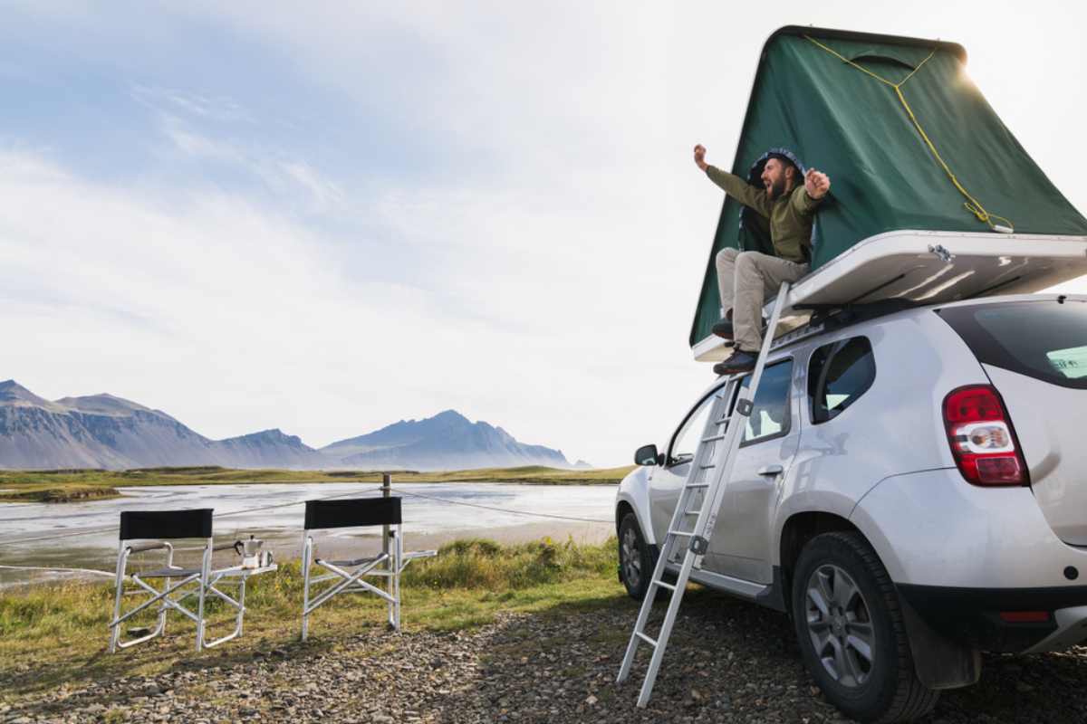 Disadvantages of renting a rooftop tent in Iceland Man stretching out from a rooftop tent on a white SUV parked by a quiet Icelandic lagoon with mountains in the background